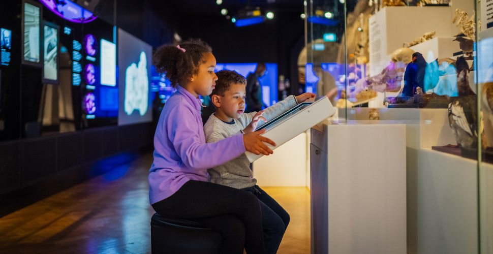 Two children explore an interactive touchscreen exhibit in a dimly lit museum gallery, with illuminated displays and artifacts behind glass.